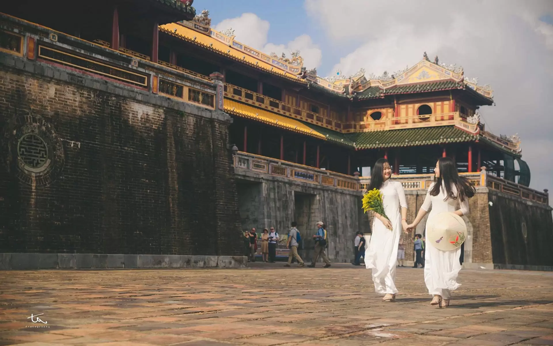 Two joyful friends in Vietnamese Ao Dai, one with a conical hat and the other with flowers, strolling in sunny Hue's imperial city in September.