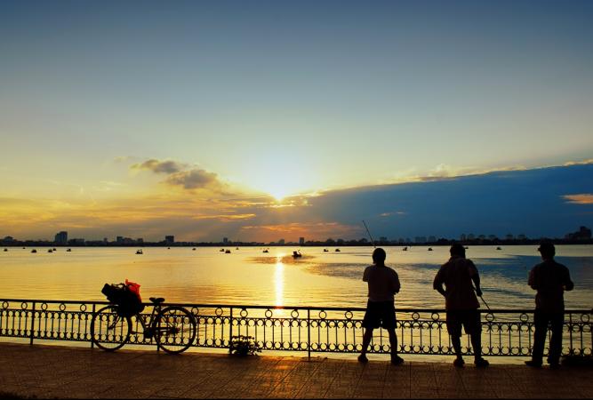 West Lake in Hanoi at sunset in July, orange sky reflected on the calm water, cityscape in the background.