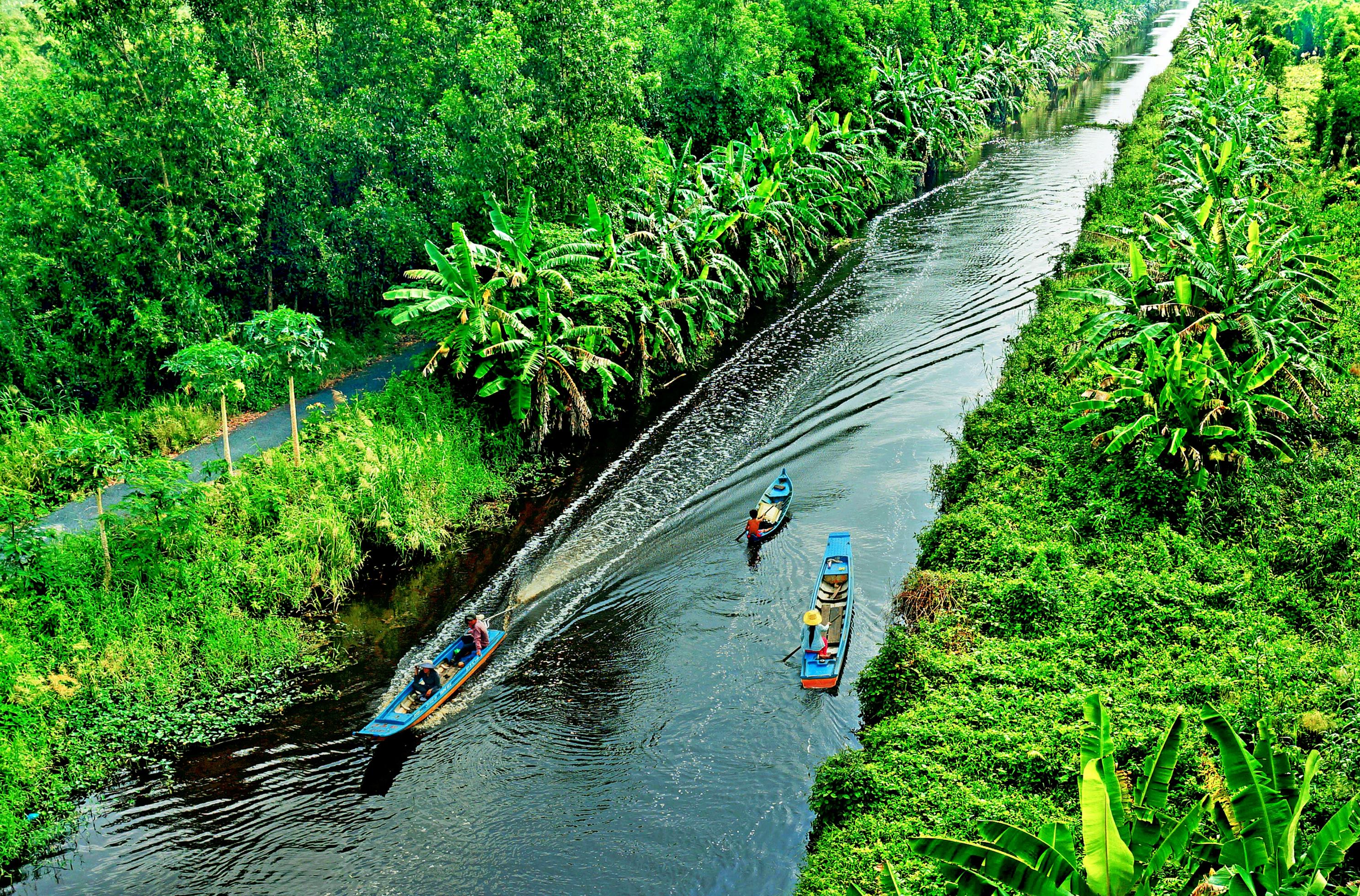 A small river next to a road, surrounded by trees in Ca Mau, Vietnam with two boats moving on the water.